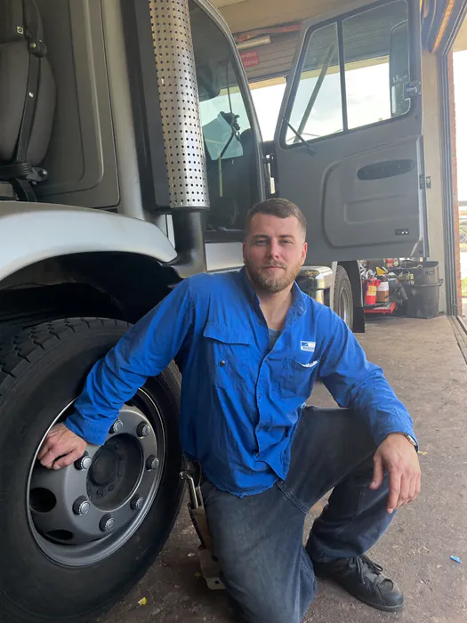 A mechanic kneeling beside the wheel of a semi truck in a shop bay